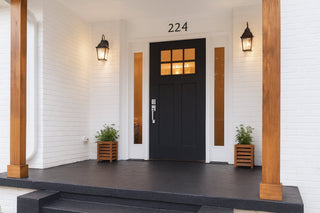 A modern house entrance featuring a black door with glass panels, white brick walls, wooden columns, and decorative lighting.